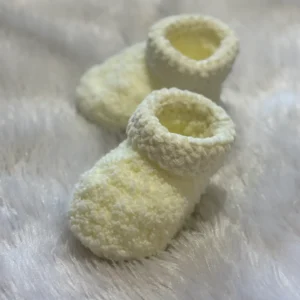 Handmade cream crochet baby booties with a soft textured stitch and ankle-height design, placed on a fluffy white background, highlighting a cozy, warm, and premium winter baby essential.