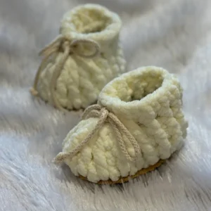 Cream-colored handmade crochet baby booties with soft textured yarn and adjustable tie strings, placed on a plush white fur background, showcasing cozy and premium newborn footwear.
