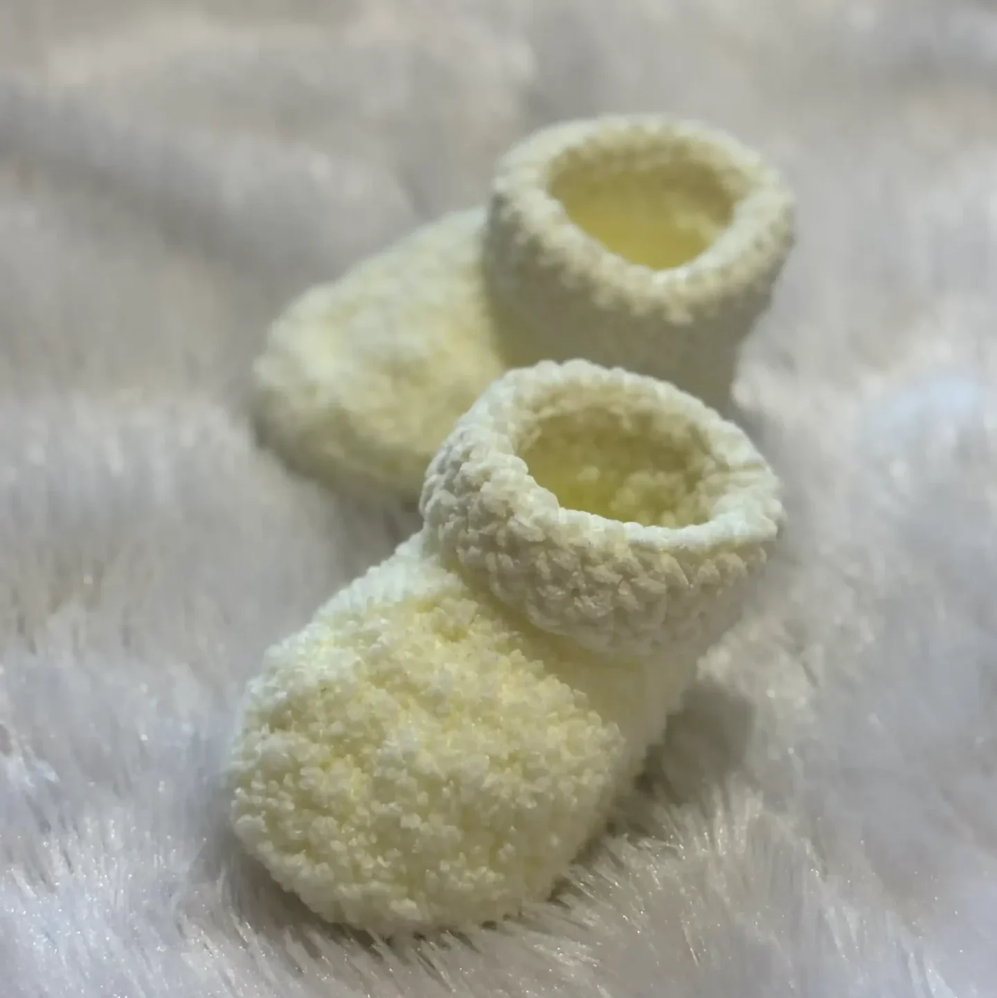 Handmade cream crochet baby booties with a soft textured stitch and ankle-height design, placed on a fluffy white background, highlighting a cozy, warm, and premium winter baby essential.