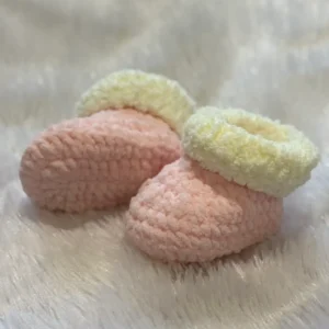 Soft, handmade crochet baby booties in blush pink with cream cuffs, placed on a plush white fabric background, showcasing cozy texture and gentle pastel tones suitable for newborns.
