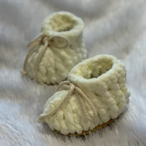 Cream-colored handmade crochet baby booties with soft textured yarn and adjustable tie strings, placed on a plush white fur background, showcasing cozy and premium newborn footwear.